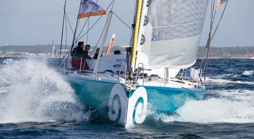 Conrad Colman's IMOCA 60 with turquoise hull and white spiral logo punching upwind in spray off the coast