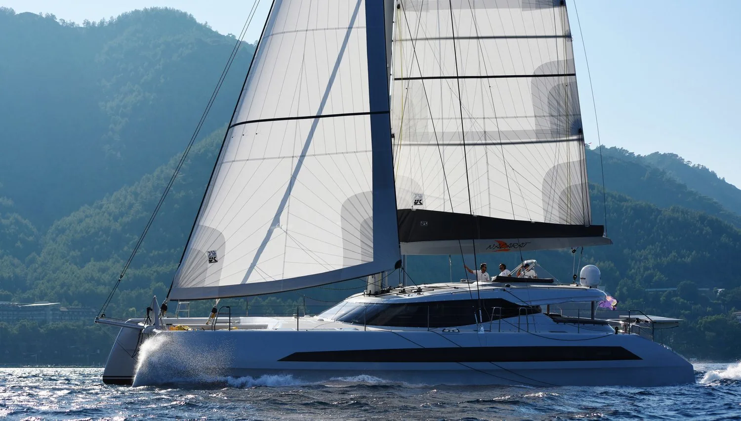 60-foot Dubois catamaran sailing under a white HydraNet Radial mainsail and genoa along a mountainous coast