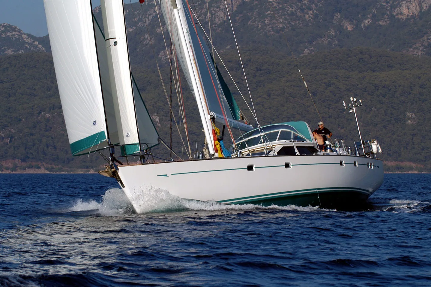 White cruising yacht with a green sheer stripe powering upwind under white sails along a mountainous coast