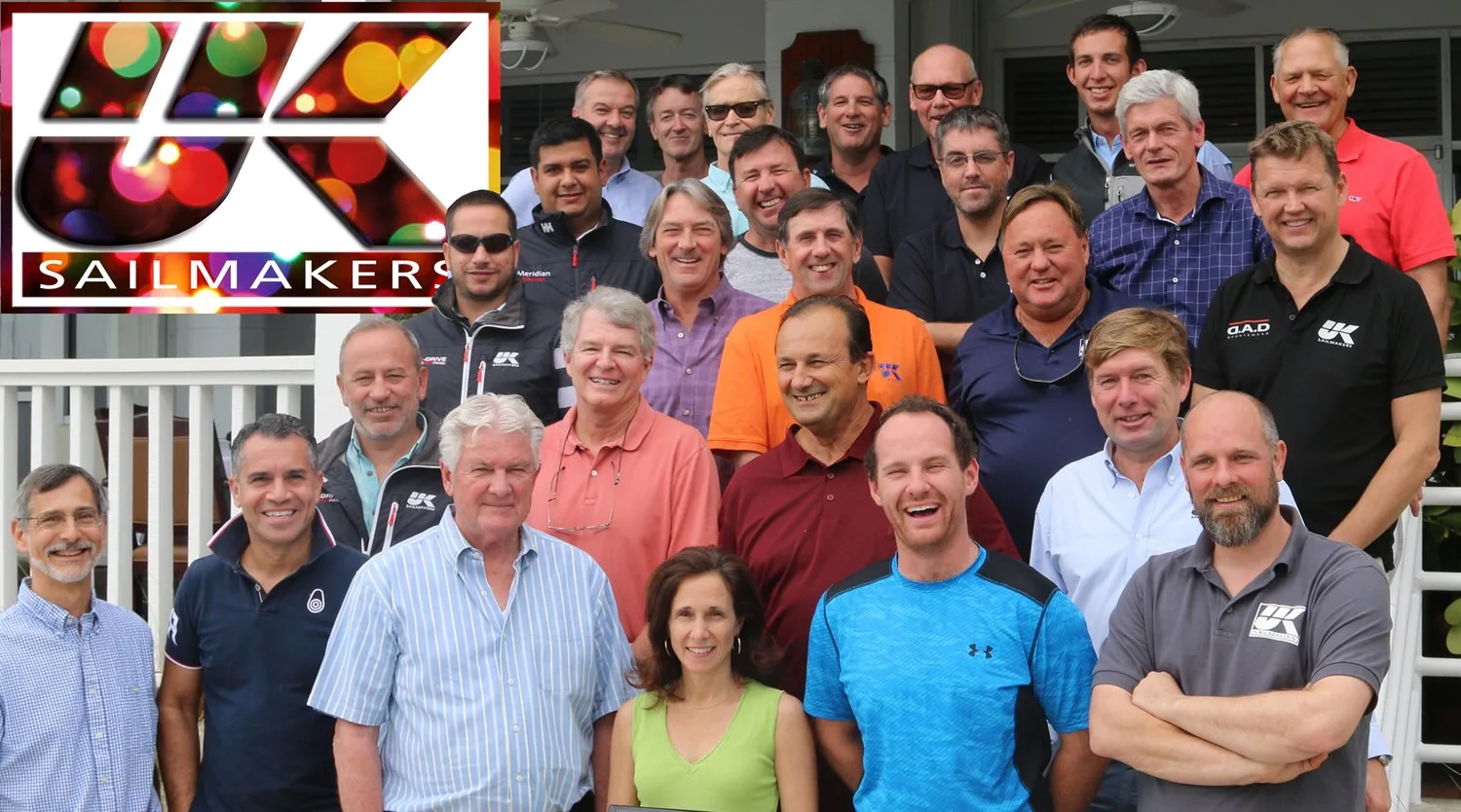 About 25 smiling UK Sailmakers team members posing outdoors, with a festive-lights UK Sailmakers logo overlaid
