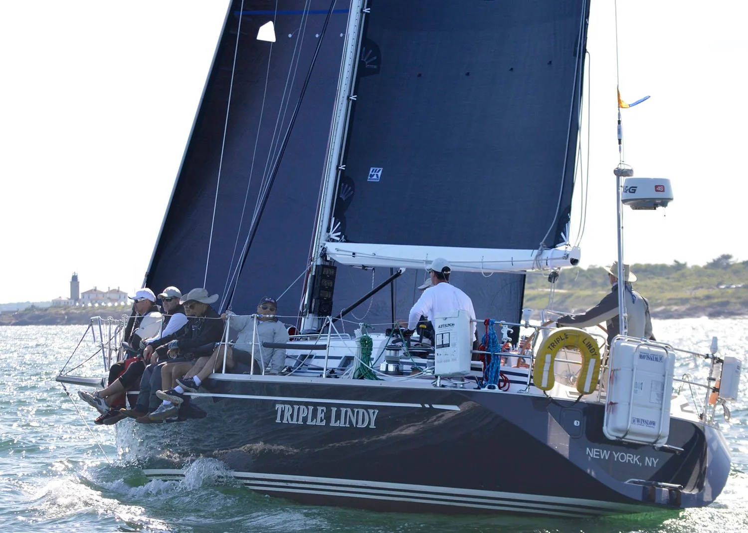 Dark-hulled TRIPLE LINDY of New York, NY sailing upwind under black sails with crew on the rail, a lighthouse astern