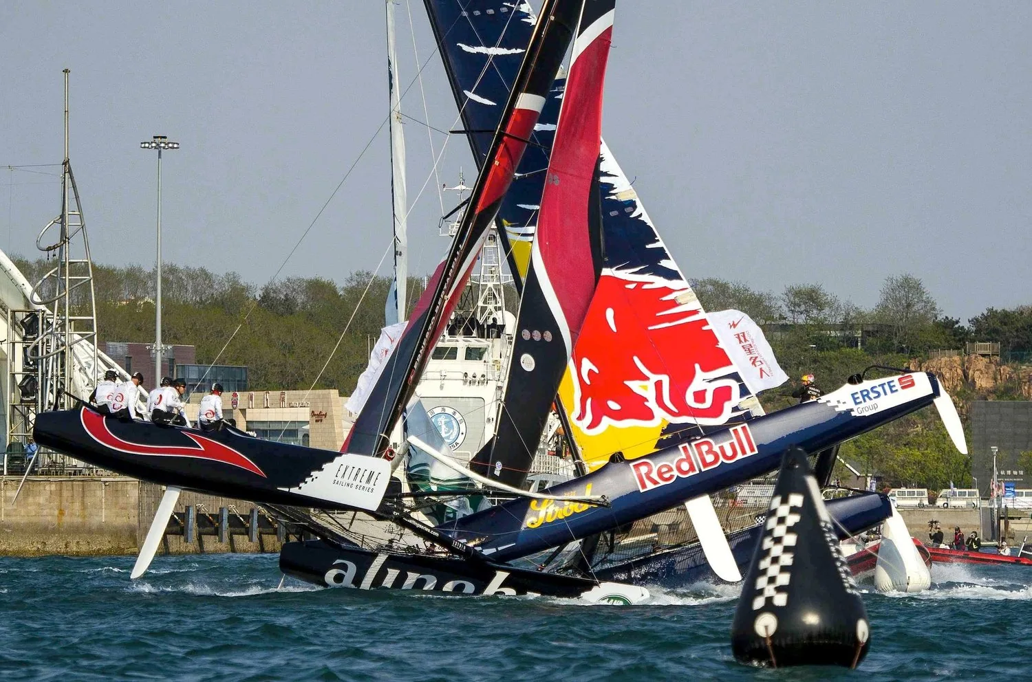 Red Bull and Alinghi racing catamarans tangled together at a black and white chequered mark in a harbour