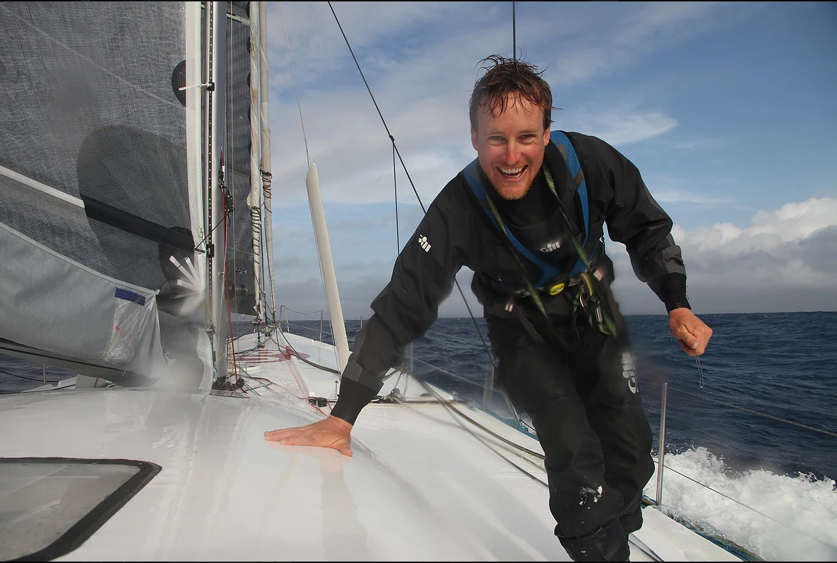 Conrad Colman grinning in a black drysuit on the wet foredeck of his IMOCA 60 at sea