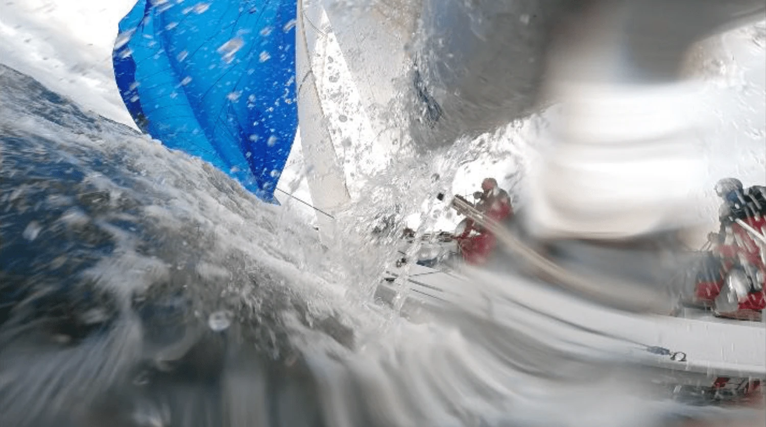 Wave spray bursting over the camera lens with a blue spinnaker and crew in red foul-weather gear behind