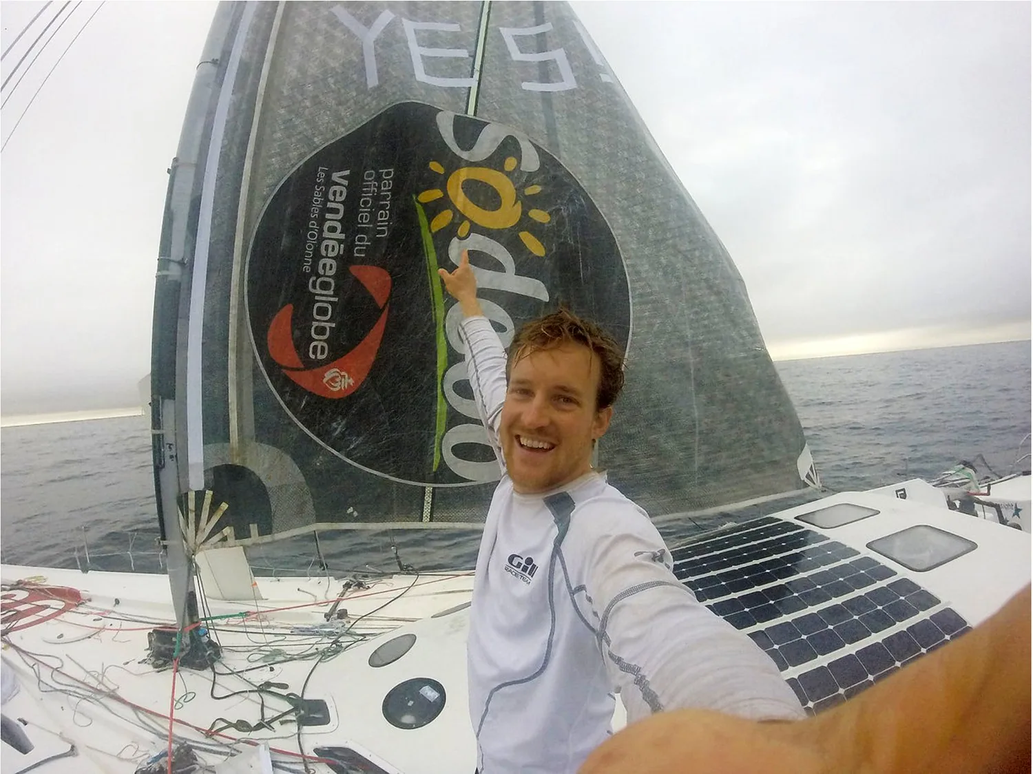 Conrad Colman pointing at his jury-rig sail marked 'YES!' with Vendée Globe logos, solar panels on the deck
