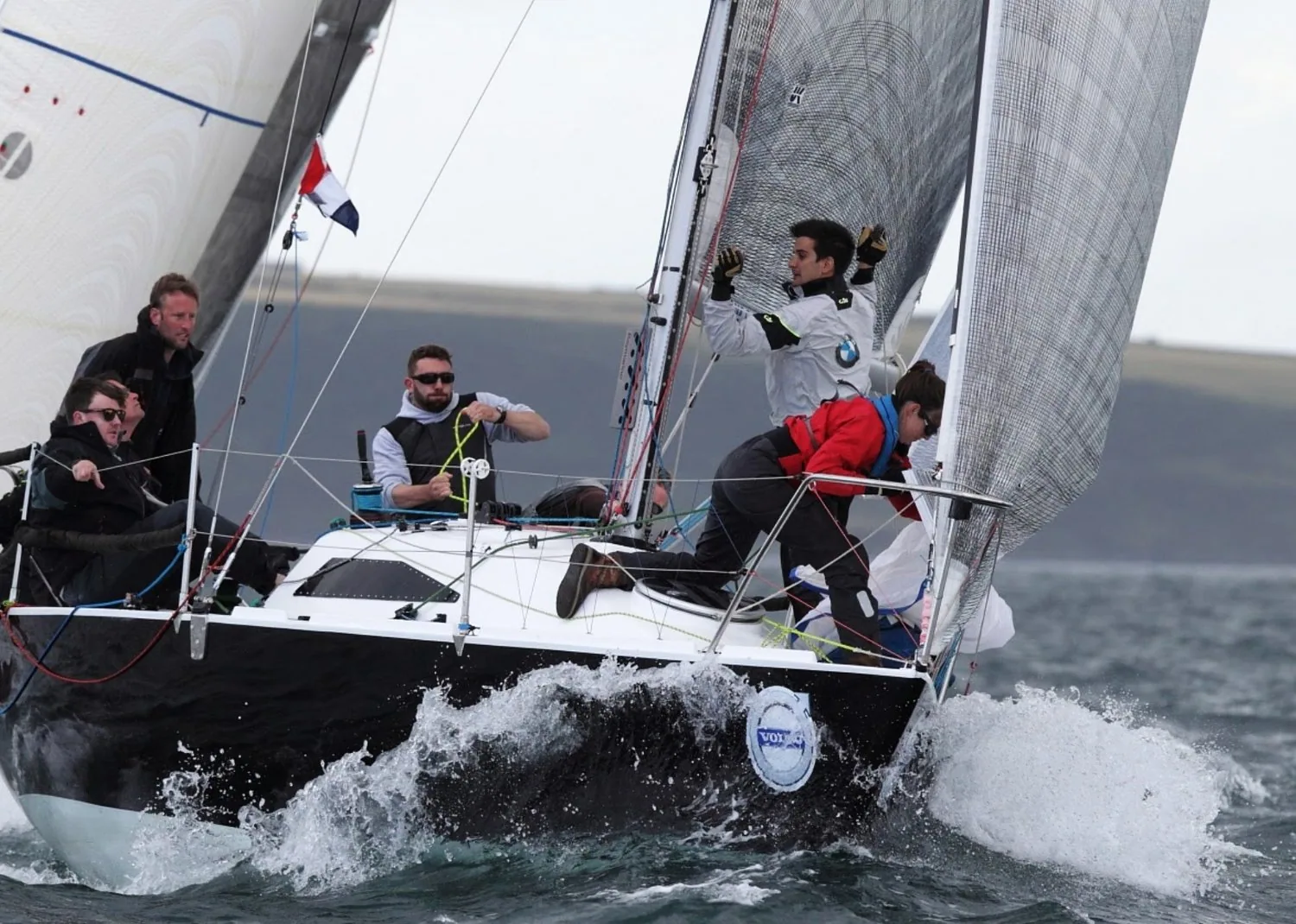 Dark-hulled sportboat crashing through a wave with five crew, one standing at the mast, another boat close astern