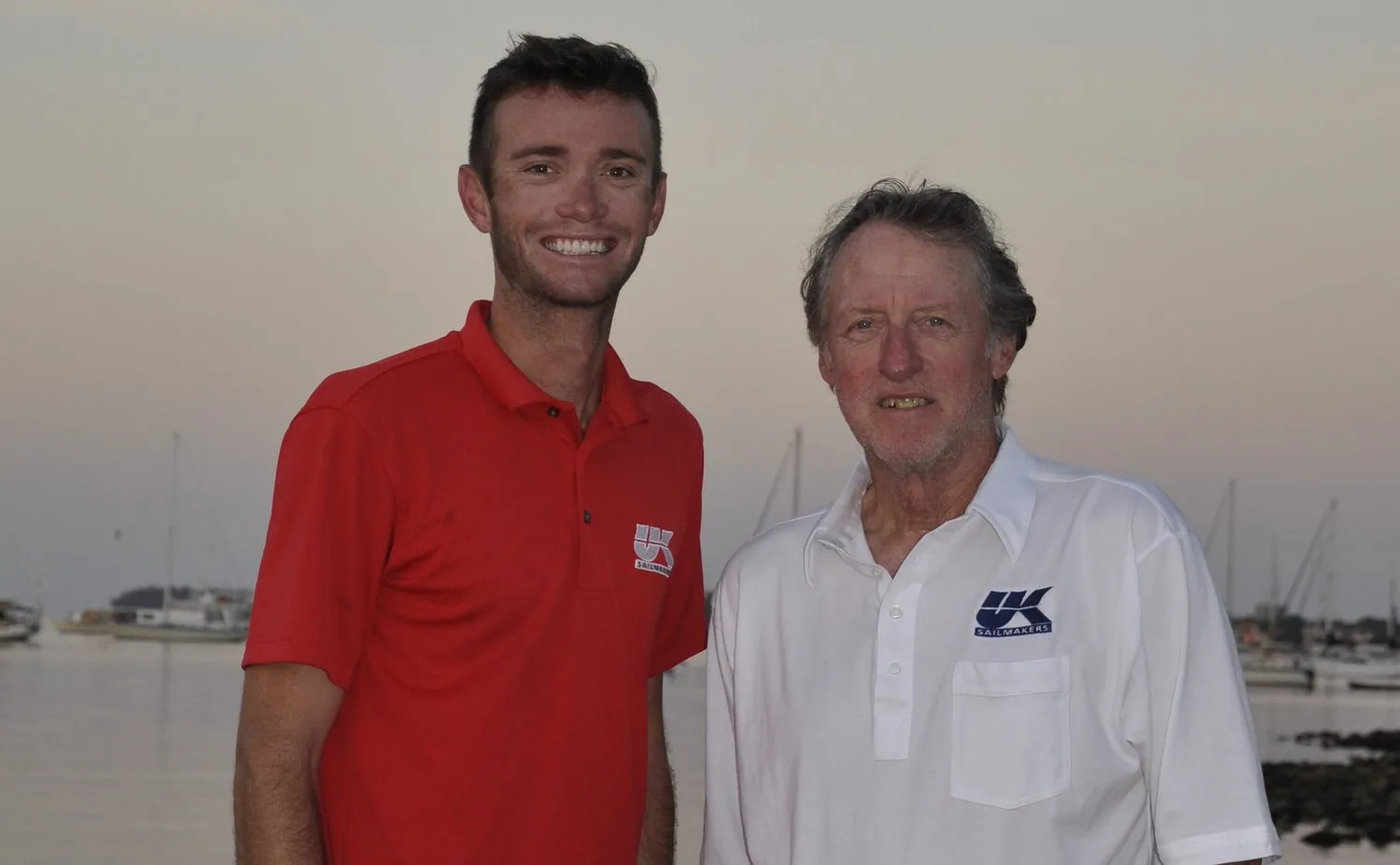 Dalton Tebo in a red polo and Ken Tebo in a white UK Sailmakers polo standing beside a harbour at dusk