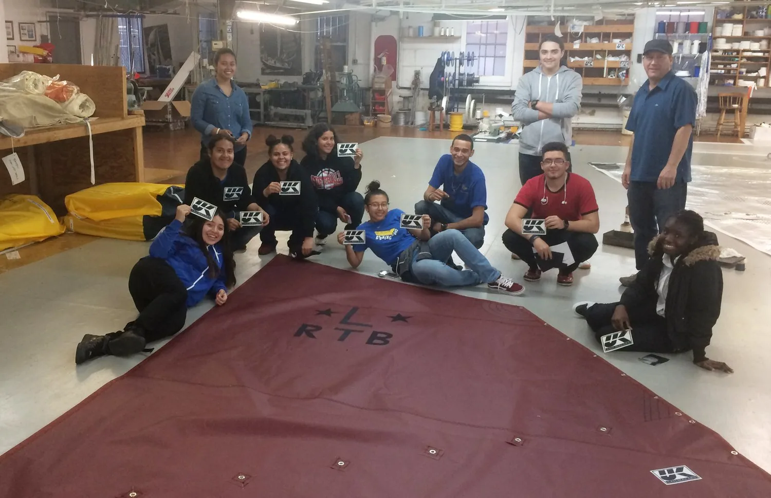 Students and mentors posing around a maroon sail lettered RTB in a sail loft, holding UK Sailmakers logo cards