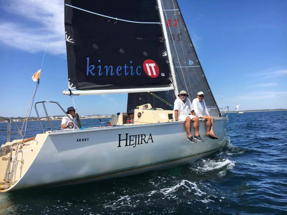 S97 HEJIRA with three crew on the rail, black kinetic IT mainsail and white hull, on a sunny bay