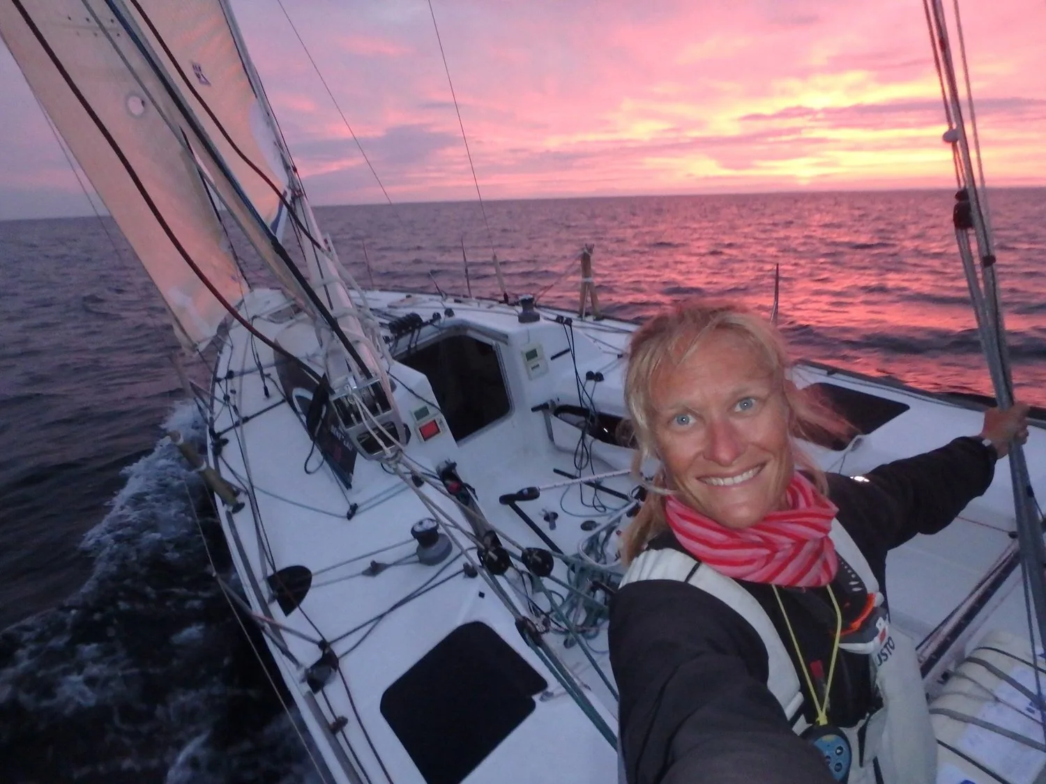 Kristen Songe Moller smiling in a selfie on her boat's foredeck under a pink sunset sky at sea