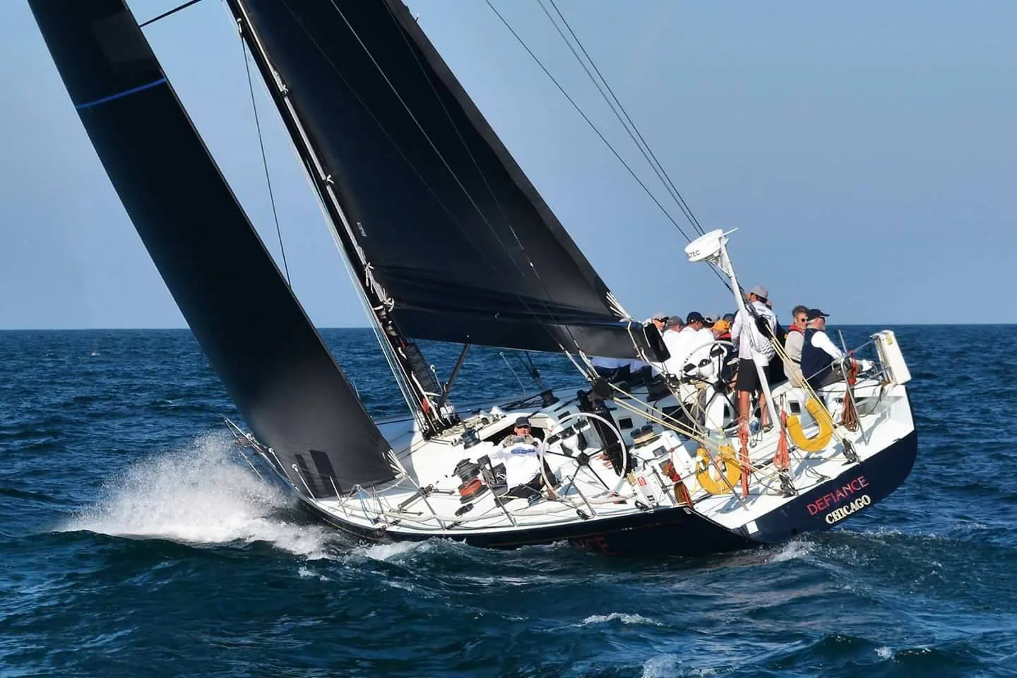 Black-hulled racing yacht DEFIANCE heeled upwind under black sails, crew on the rail, spray off the bow