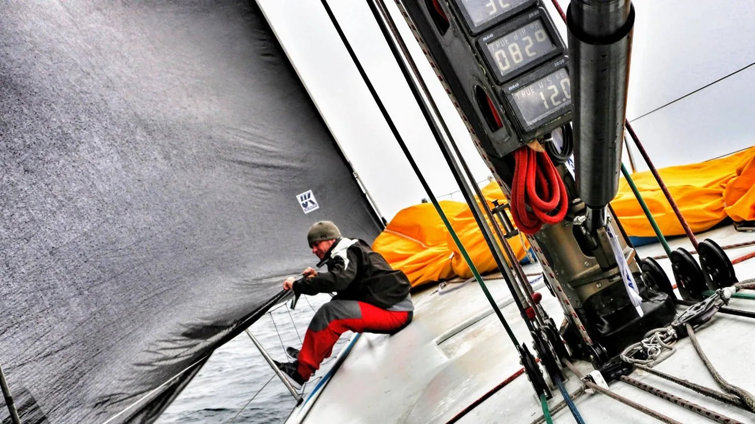 Bowman in red foul-weather gear crouched at the bow of DEFIANCE beside a grey Uni-Titanium headsail and orange sail bags