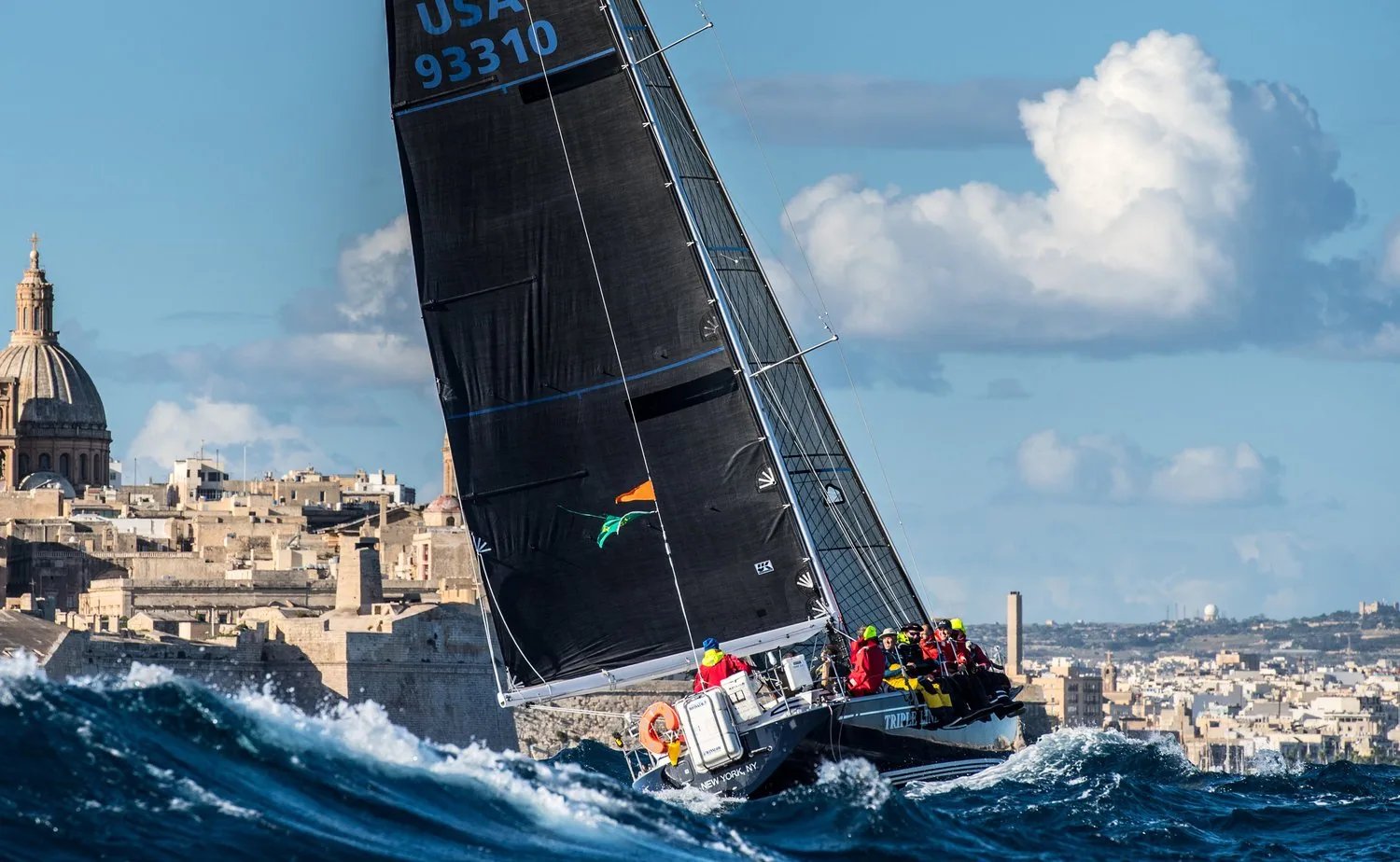 TRIPLE LINDY, USA 93310, black sails, crashing through blue swell off Malta's domed skyline in the Middle Sea Race