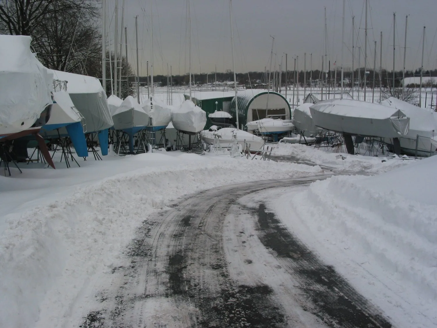Boatyard buried in snow: rows of covered sailboats on stands under bare masts, a ploughed track winding through