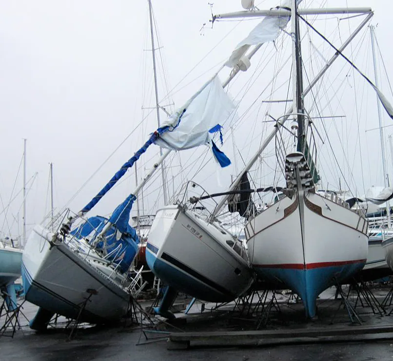 Sailboats toppled off their stands in a winter boatyard, one mast leaning with a shredded blue cover flapping