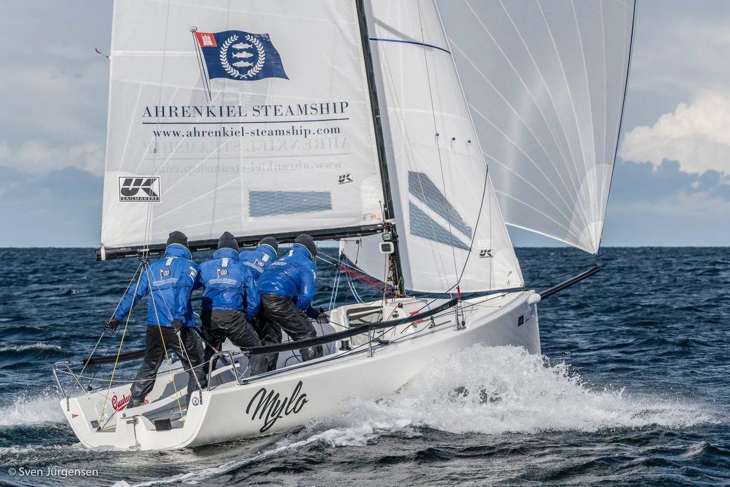 J/70 MYLO planing downwind under a white asymmetric spinnaker, four crew in blue hiking, sponsor logo on the mainsail