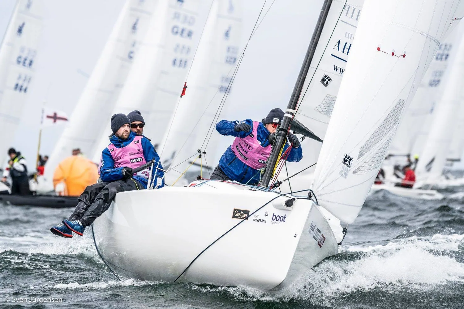 Bow-on view of a J/70 beating upwind through spray, three crew in pink bibs hiking, a packed fleet astern