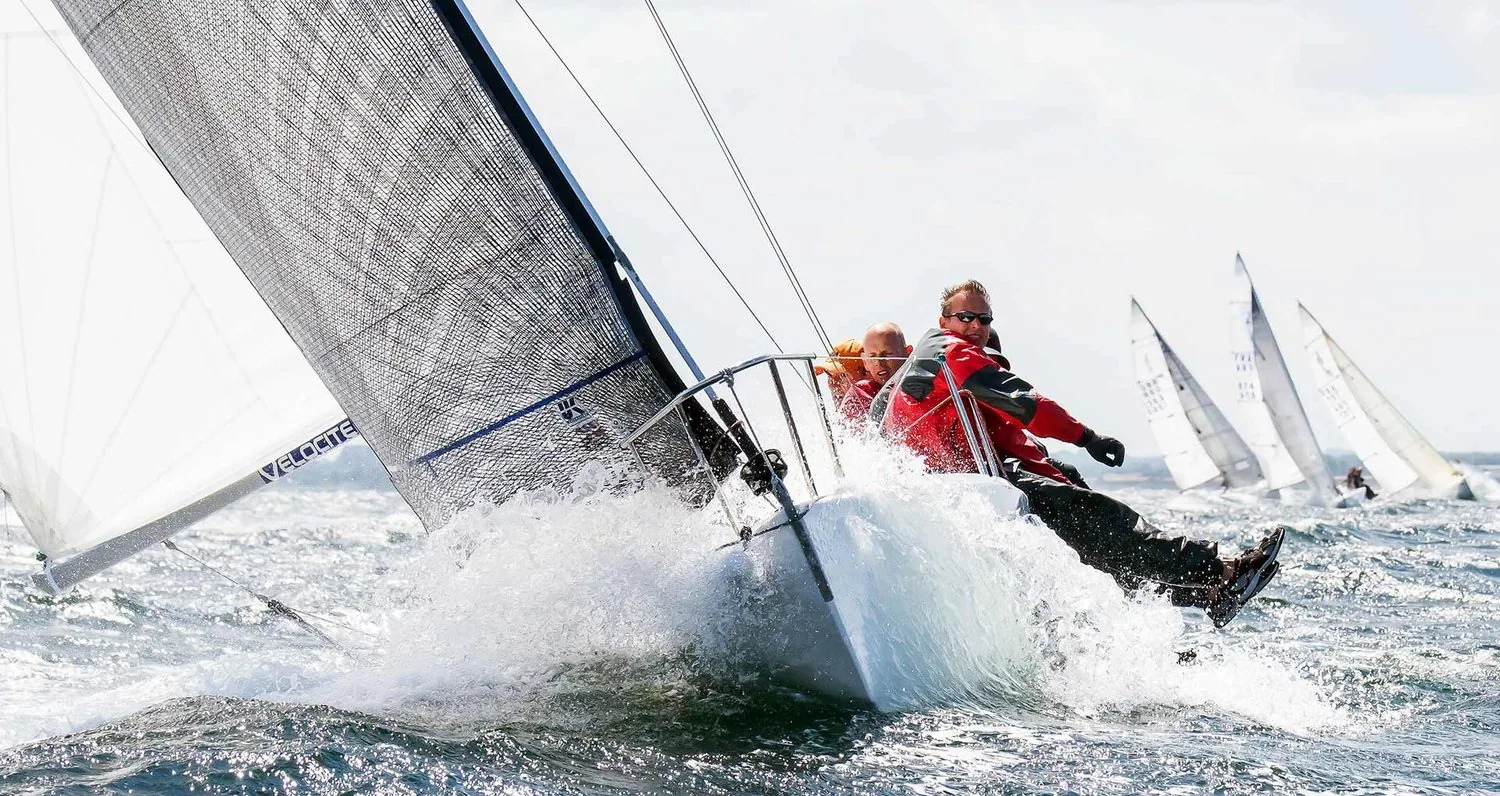 J/80 PIKE surfing down a wave in big breeze under a grey mainsail and white spinnaker, crew in red hiking aft