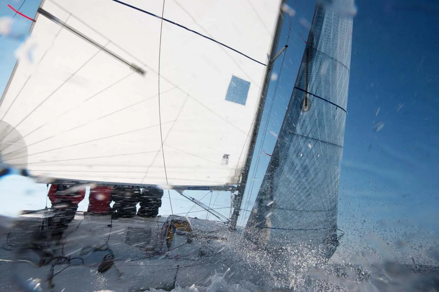 J/80 heeled hard upwind through spray, white mainsail and jib, crew in red hiking, shot through a water-spattered lens