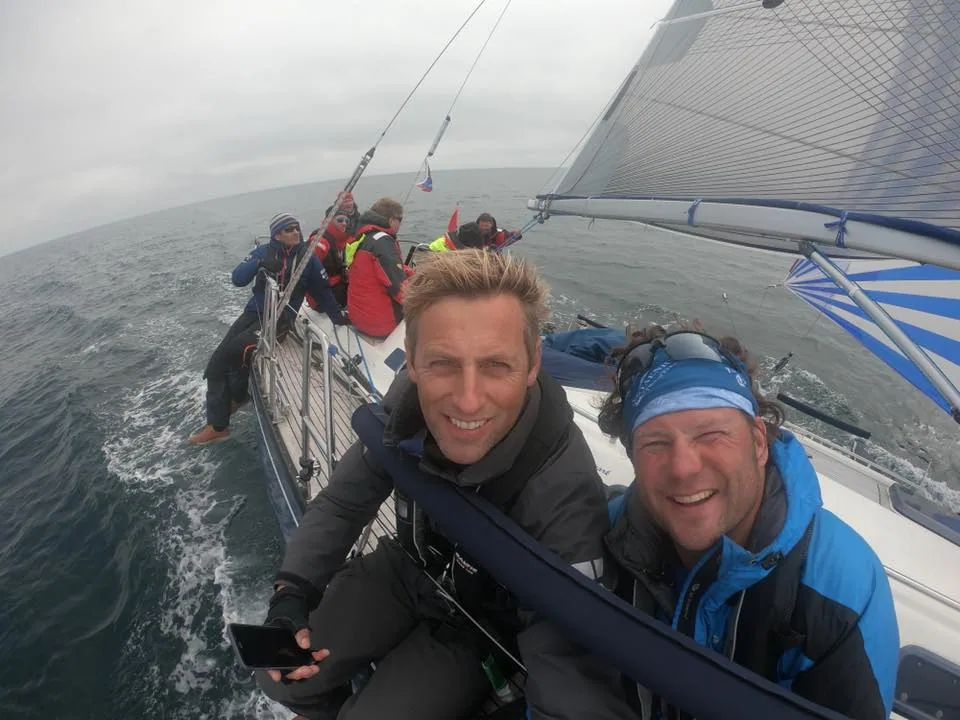 Two grinning crew in foul-weather gear take a wide-angle selfie at the stern, crewmates on the rail and grey sea behind