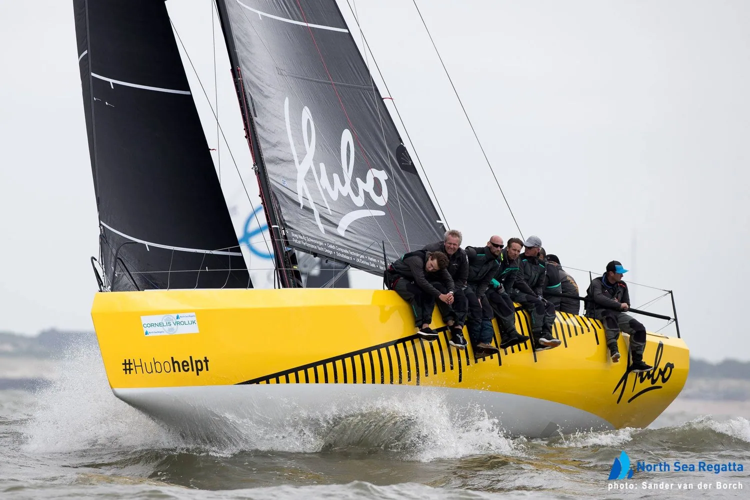 Yellow Waarschip 36 HUBO heeled upwind under black sails, crew of seven hiking on the rail, #Hubohelpt on the hull