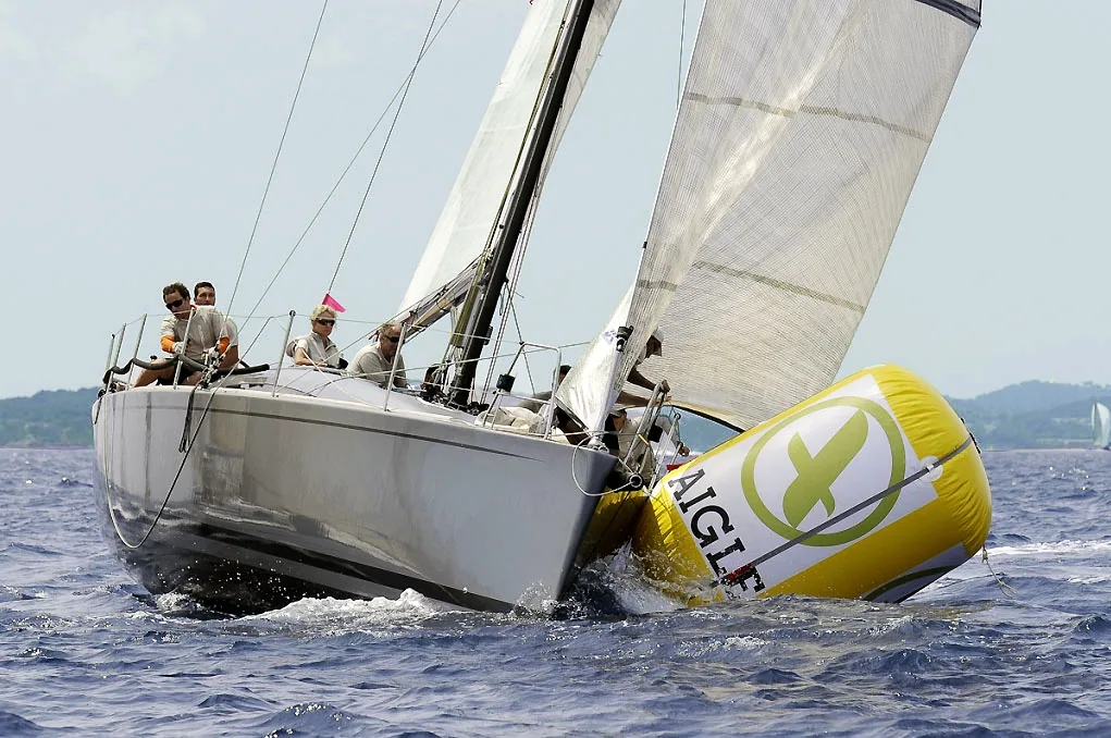 Grey racing yacht ploughing into a yellow inflatable race mark, the mark crushed under the bow as the crew look on