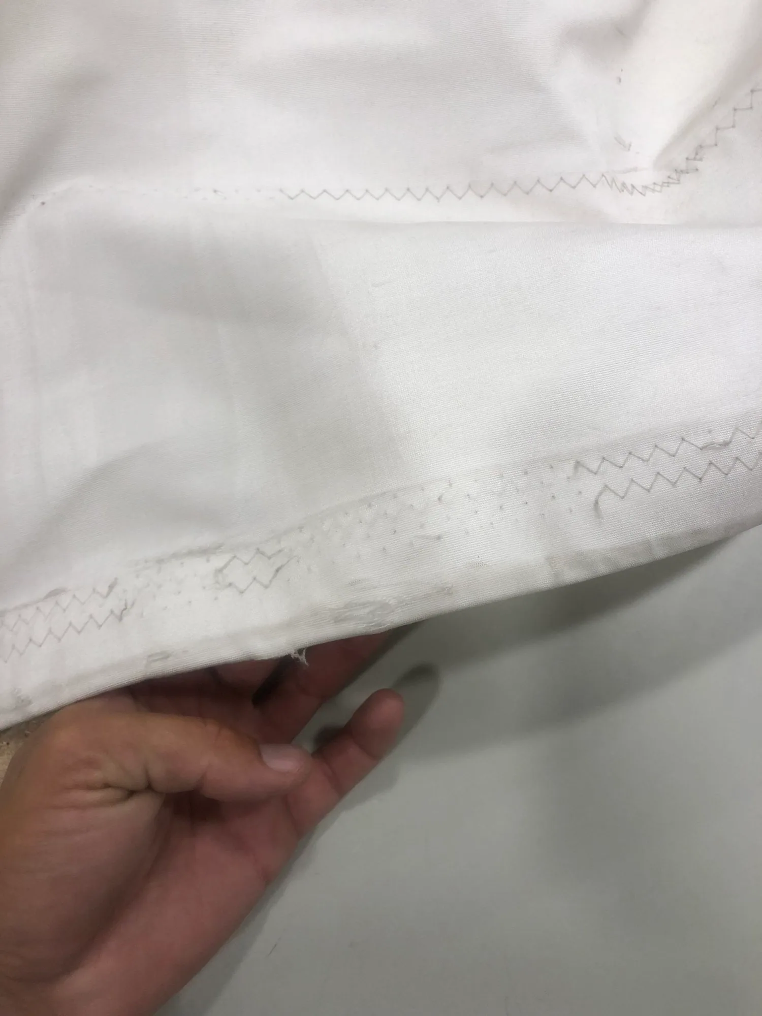 Hand holding the edge of a white sail to show chafe: worn cloth and frayed threads along a zigzag-stitched seam