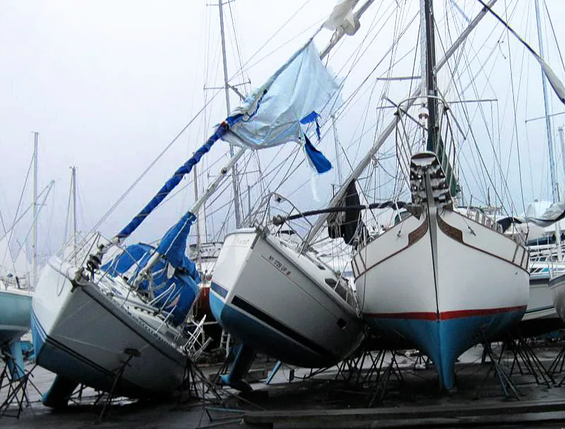 Sailboats knocked off their stands in a winter boatyard, a leaning mast trailing a torn blue cover