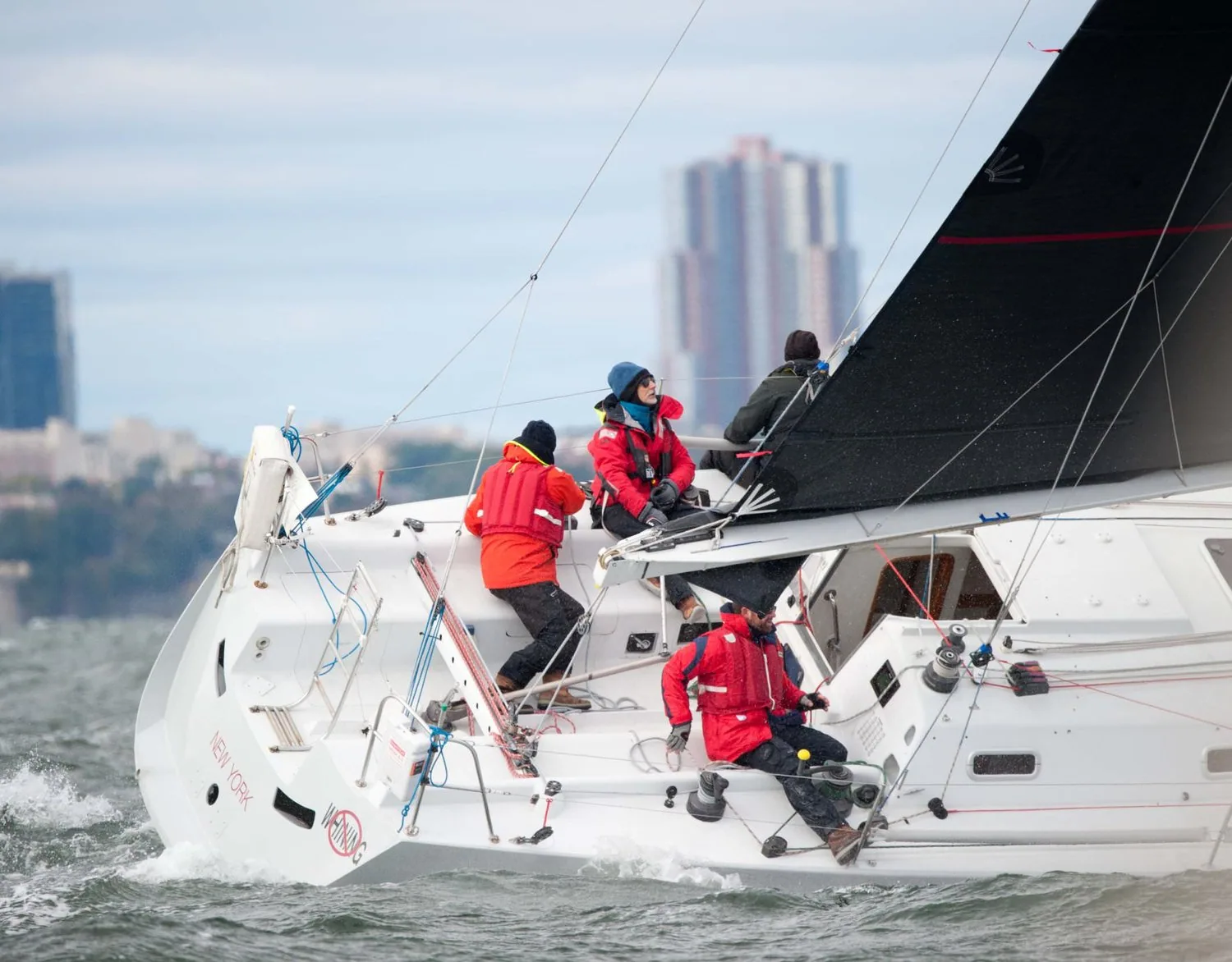 Three crew in red foul-weather gear on a white racer-cruiser with a black mainsail, bowman on the pole, city towers behind