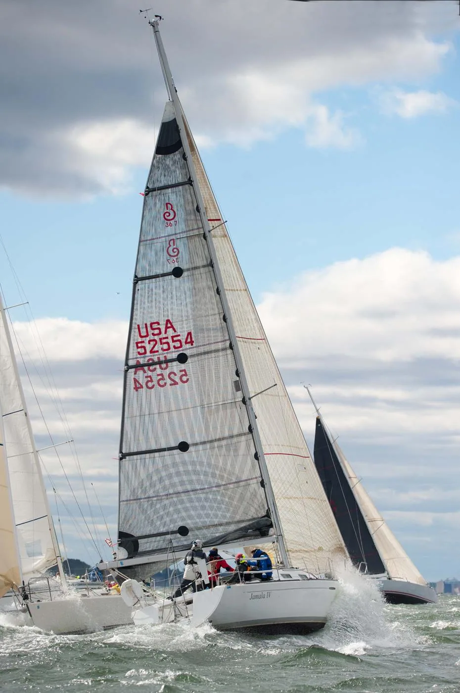Racer USA 52554 heeled upwind under a grey laminate mainsail in choppy water, crew in red, rival yachts on both sides