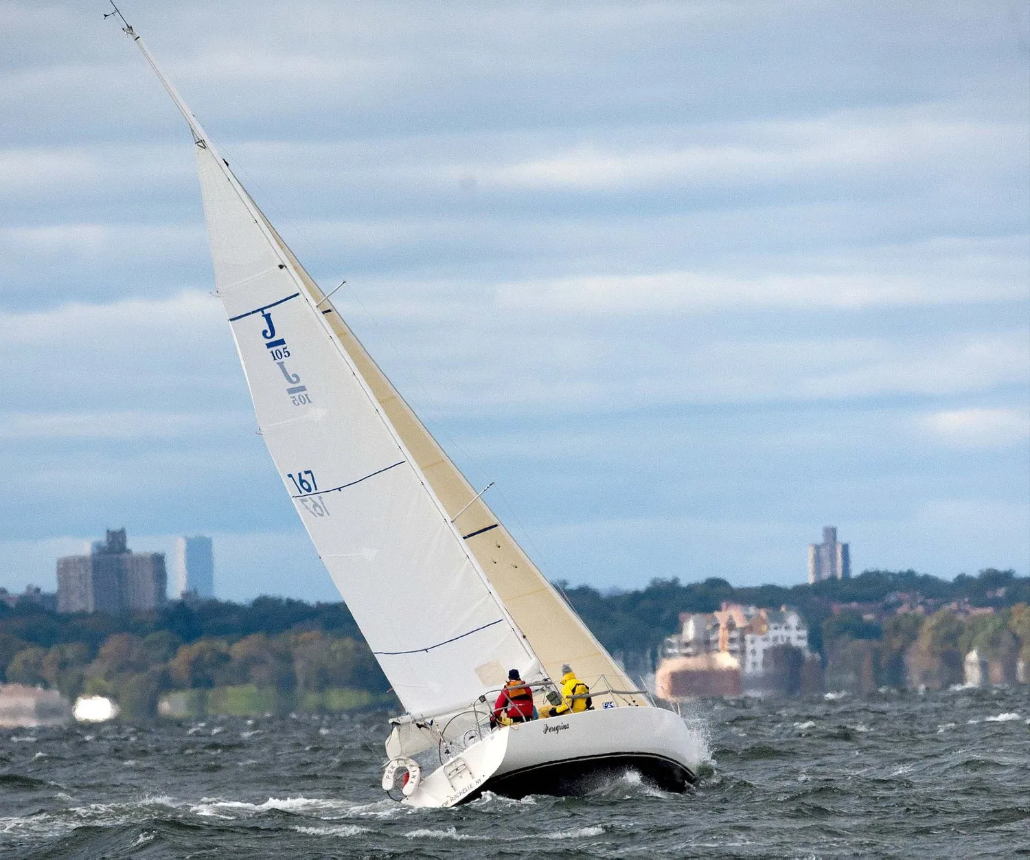 J/105 number 167 heeled upwind in grey chop under white sails, two crew in red and yellow, city buildings on the shore
