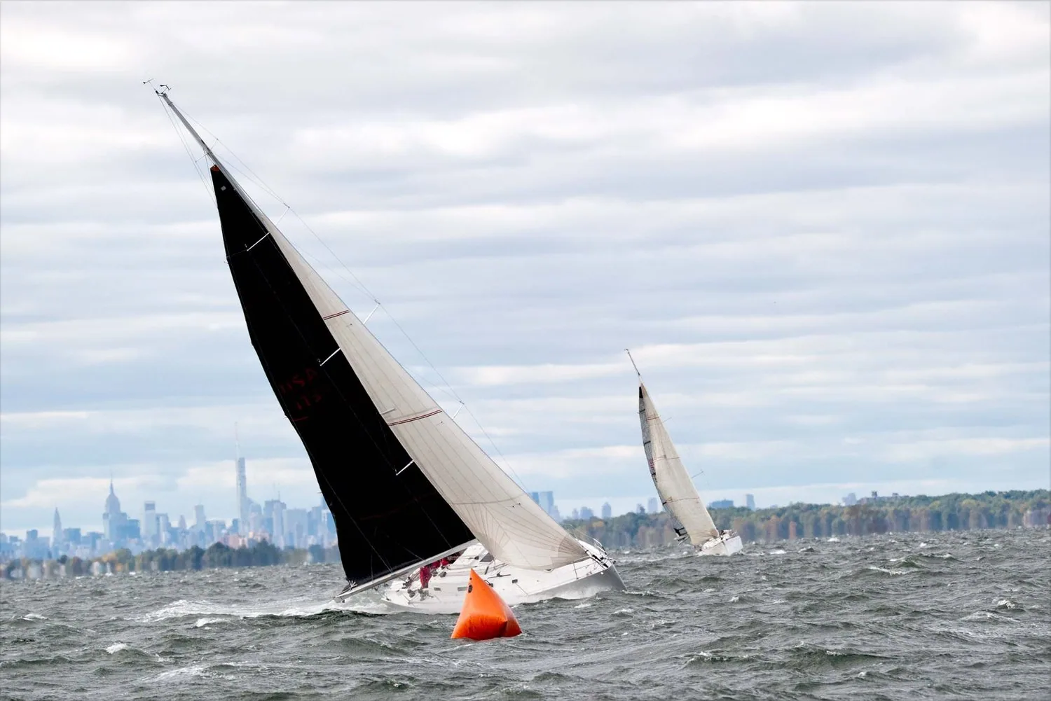 Two yachts beating past an orange inflatable mark in grey chop, the nearer with a black mainsail, city skyline on the horizon