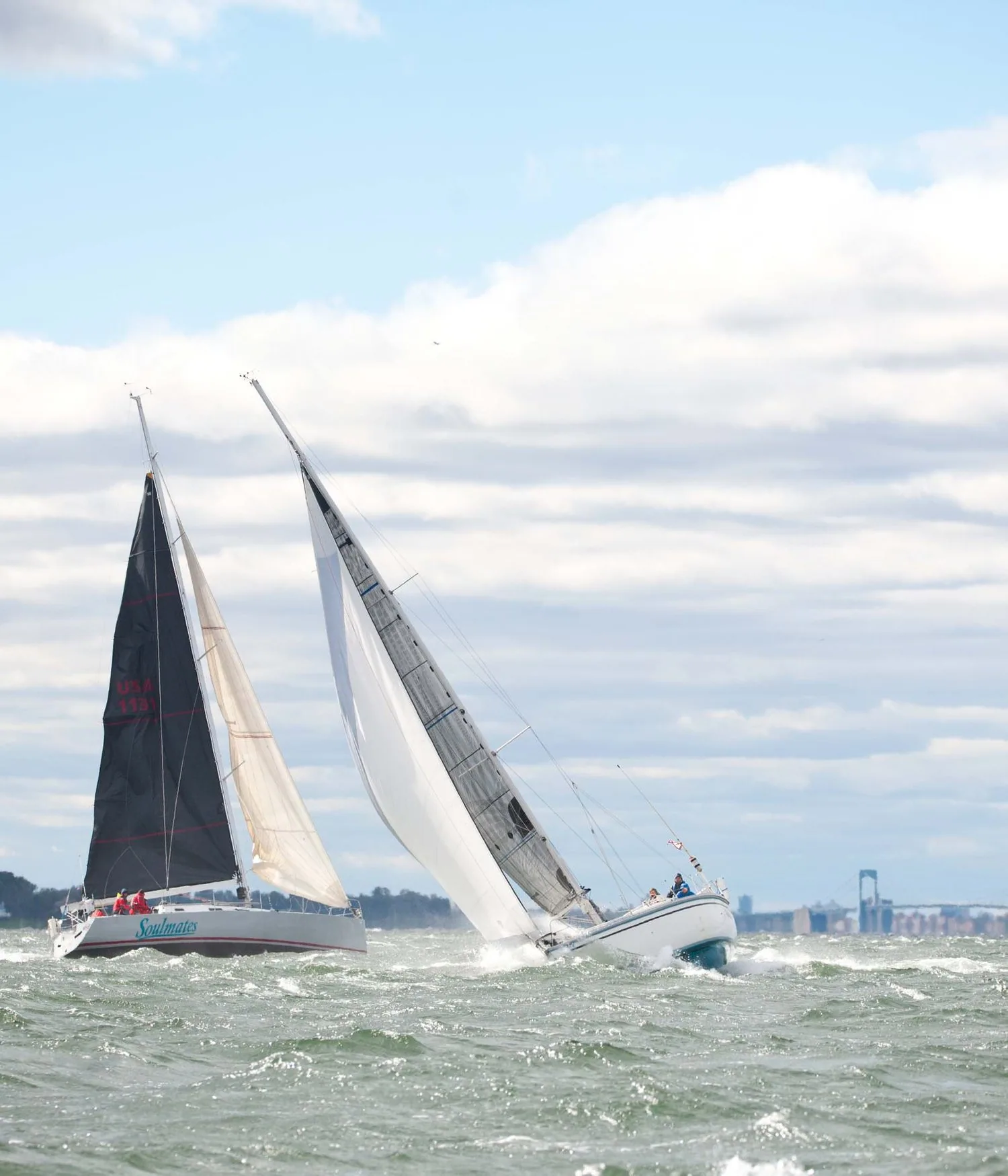 Two yachts heeled hard upwind in choppy water, one with a black mainsail, one all-white, a suspension bridge on the horizon