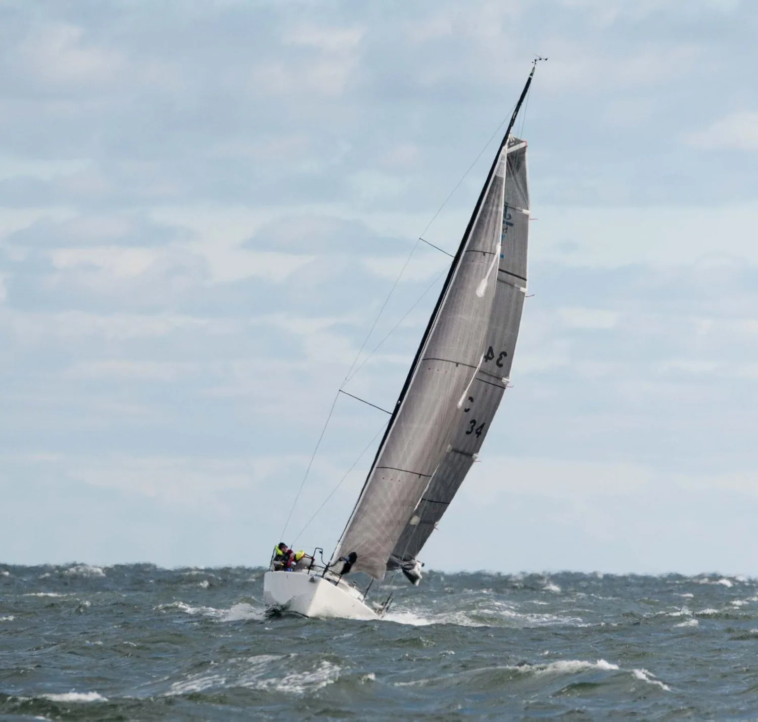 Small racer heeled hard upwind under a grey laminate mainsail, sail number 34, two crew aboard in choppy water