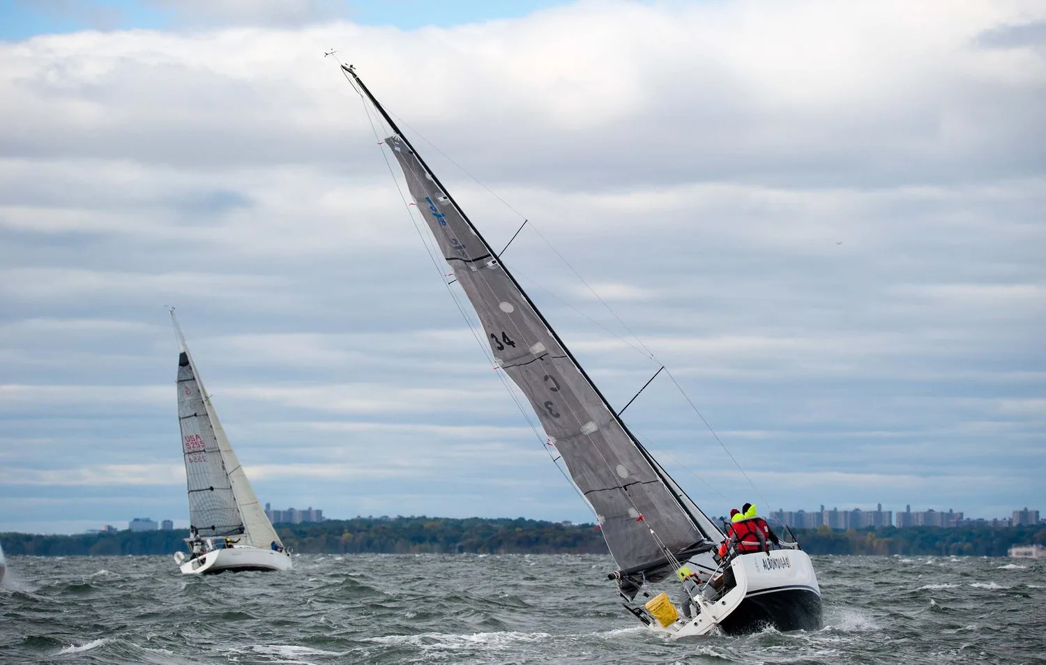 Two yachts beating upwind in grey chop, the nearer under a grey laminate mainsail with crew in yellow, wooded shore behind