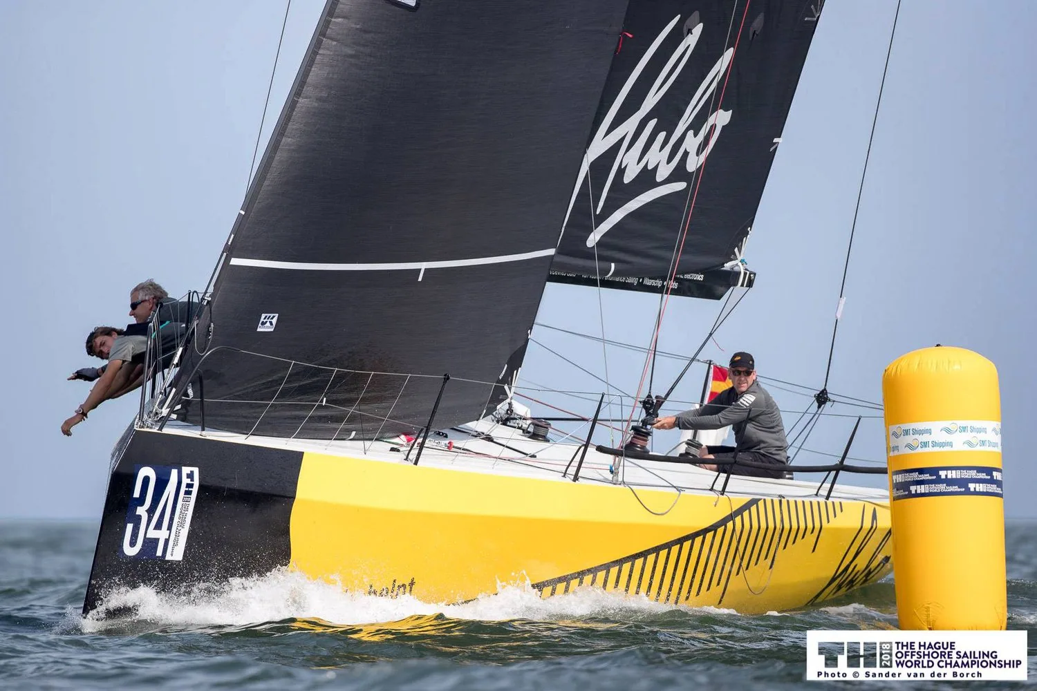 HUBO, yellow hull with bow number 34, rounding a yellow inflatable mark under black sails, crew on the rail and helmsman aft
