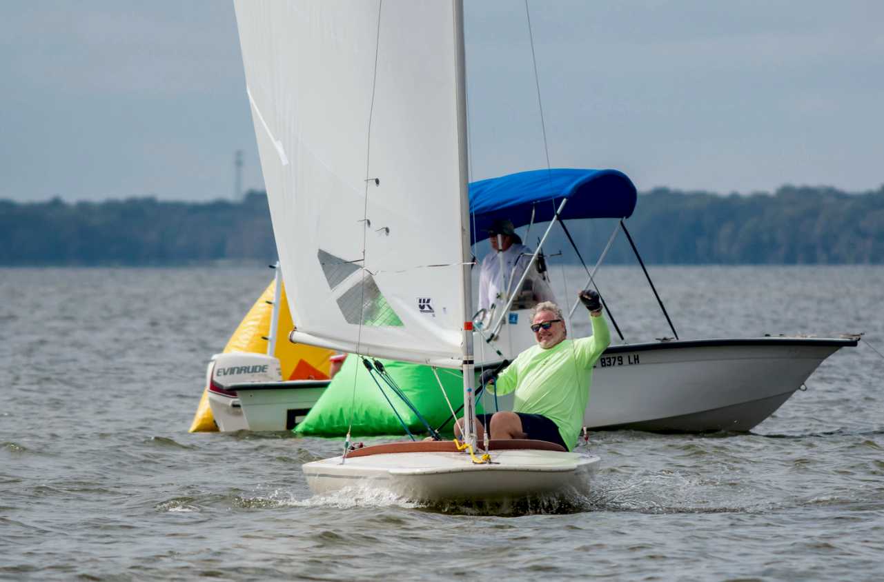 MC Scow sailor in a lime-green shirt hiking past inflatable marks, white UK Sailmakers mainsail, committee boat behind