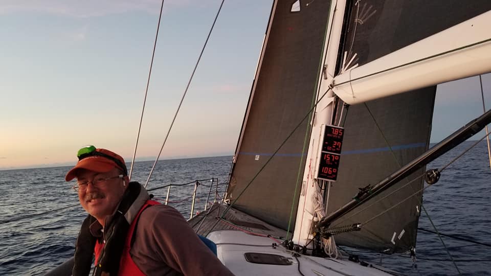 Crew member in an orange cap and red life jacket smiling on deck at dusk beneath a dark grey mainsail with mast instruments