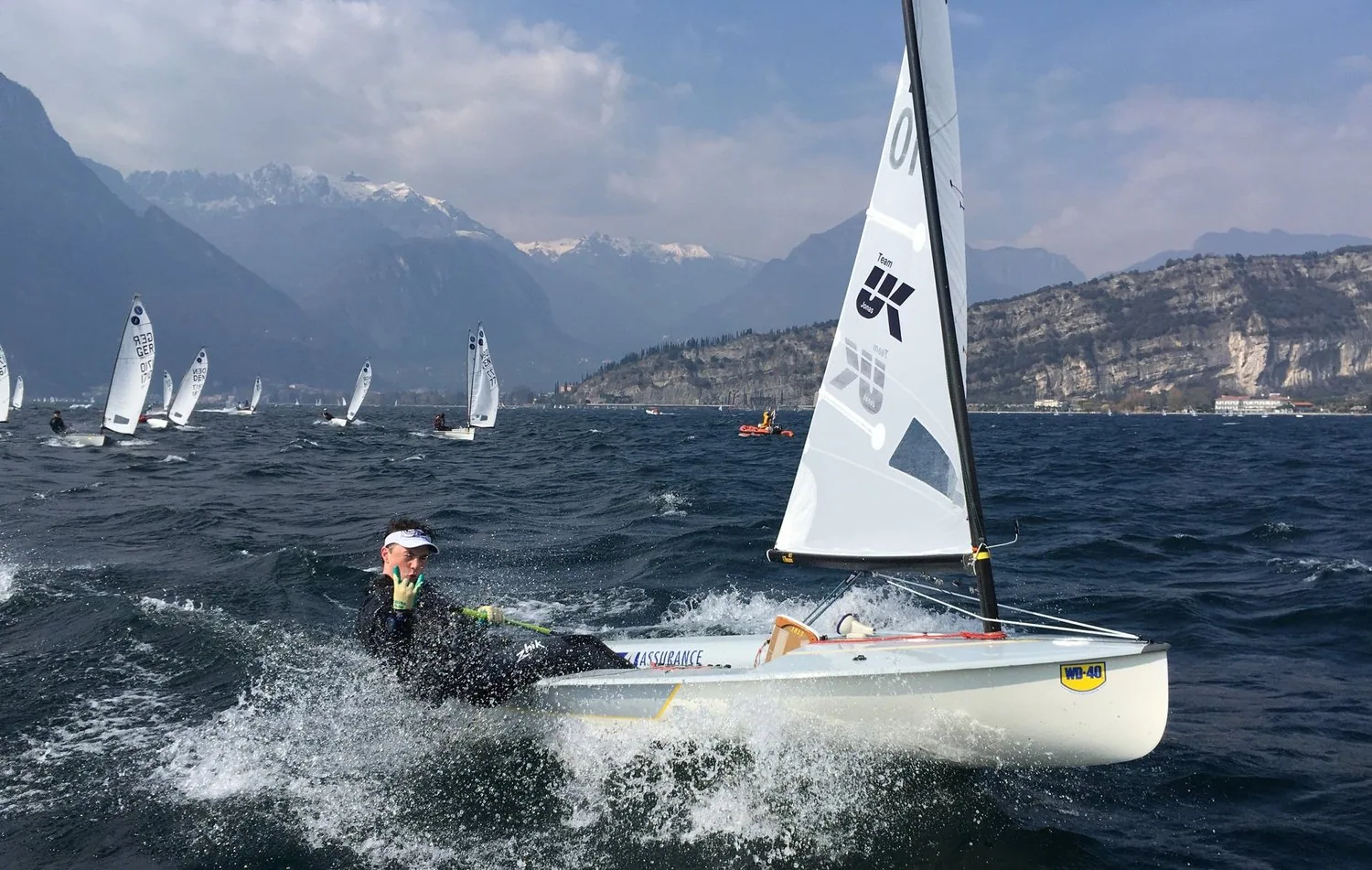 Europe dinghy planing on a mountain lake, sailor hiking hard, UK Sailmakers logo on the sail, fleet and snowy peaks behind