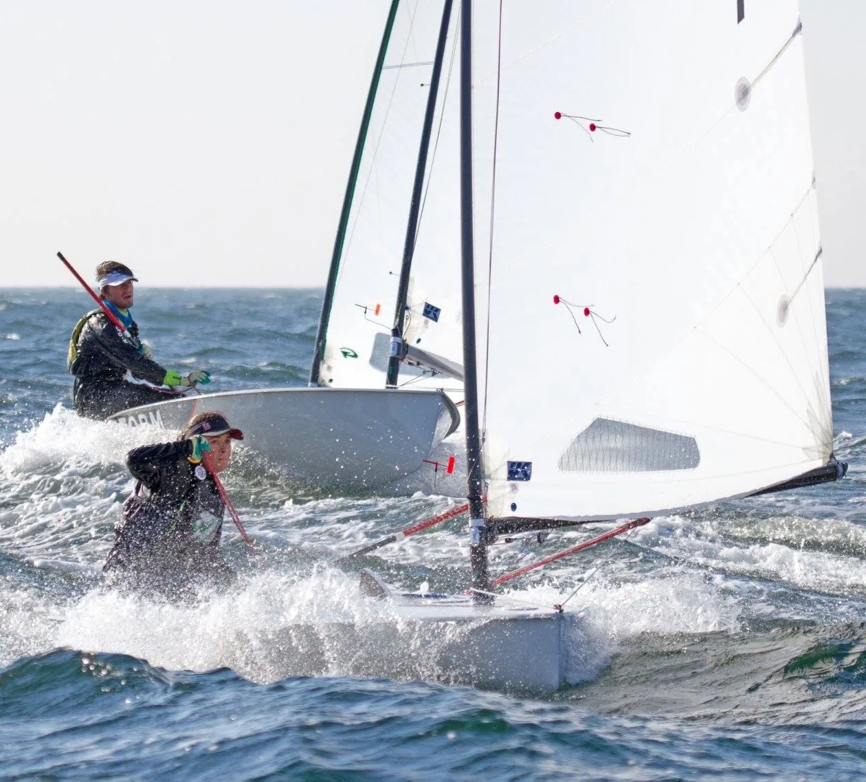 Two Europe dinghy sailors hiking hard side by side in spray, white mainsails with red telltales