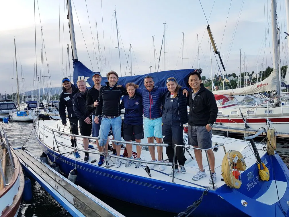 Nine crew posing on the deck of the blue-hulled IMX 38 BRAVEHEART in a marina, UK Sailmakers logo on the mainsail