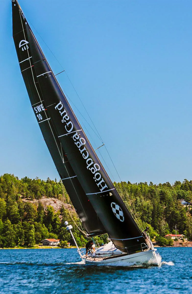 Arcona 465 FLYT heeled hard under black Högberga Gård sails, SWE 10, passing a wooded shore with red cottages