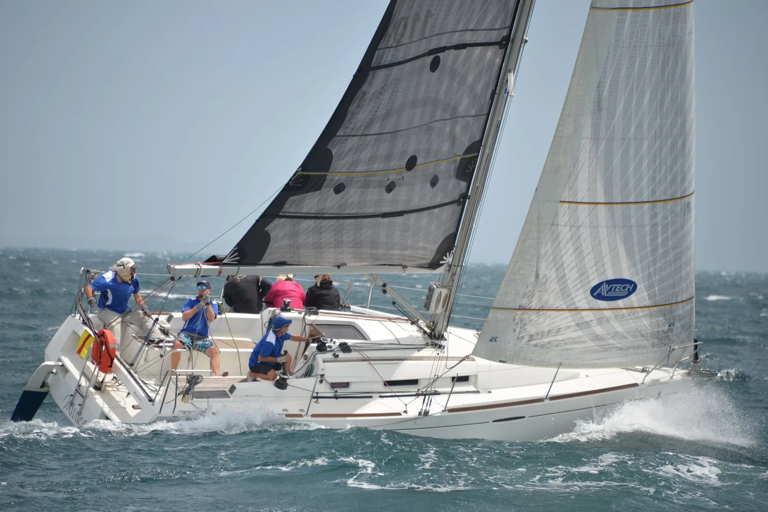 Sun Fast 32 driving upwind in spray under a dark grey mainsail and white genoa, three crew in blue at the stern
