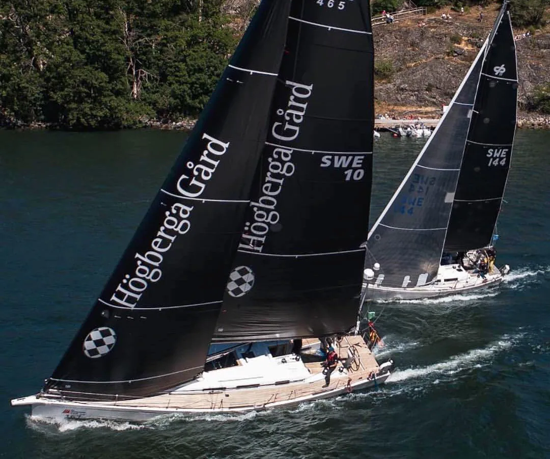 Aerial view of Arcona 465 FLYT under black Högberga Gård sails leading SWE 144 along a rocky wooded shoreline