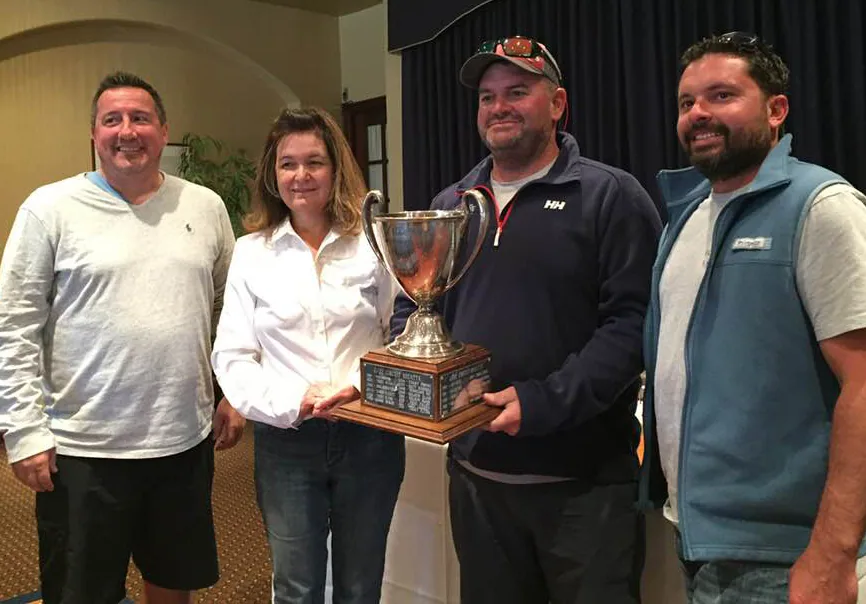 Four people holding a large silver trophy cup on an engraved wooden base at an indoor prize-giving