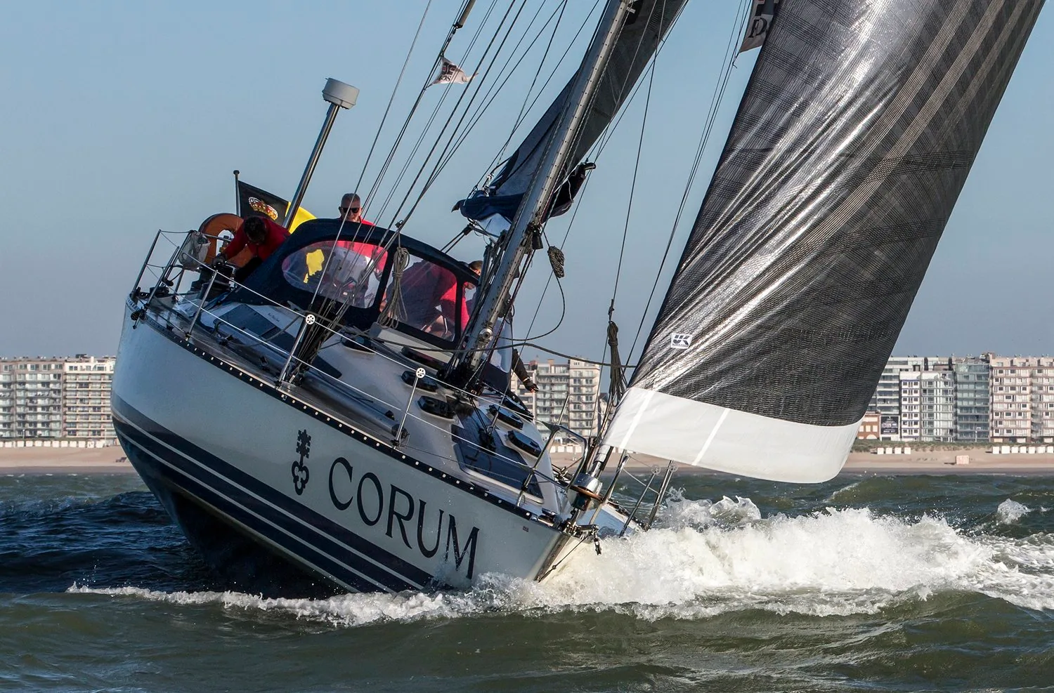 X-452 NINIX heeled hard under grey X-Drive® sails, CORUM lettering on the hull, seaside apartment blocks behind