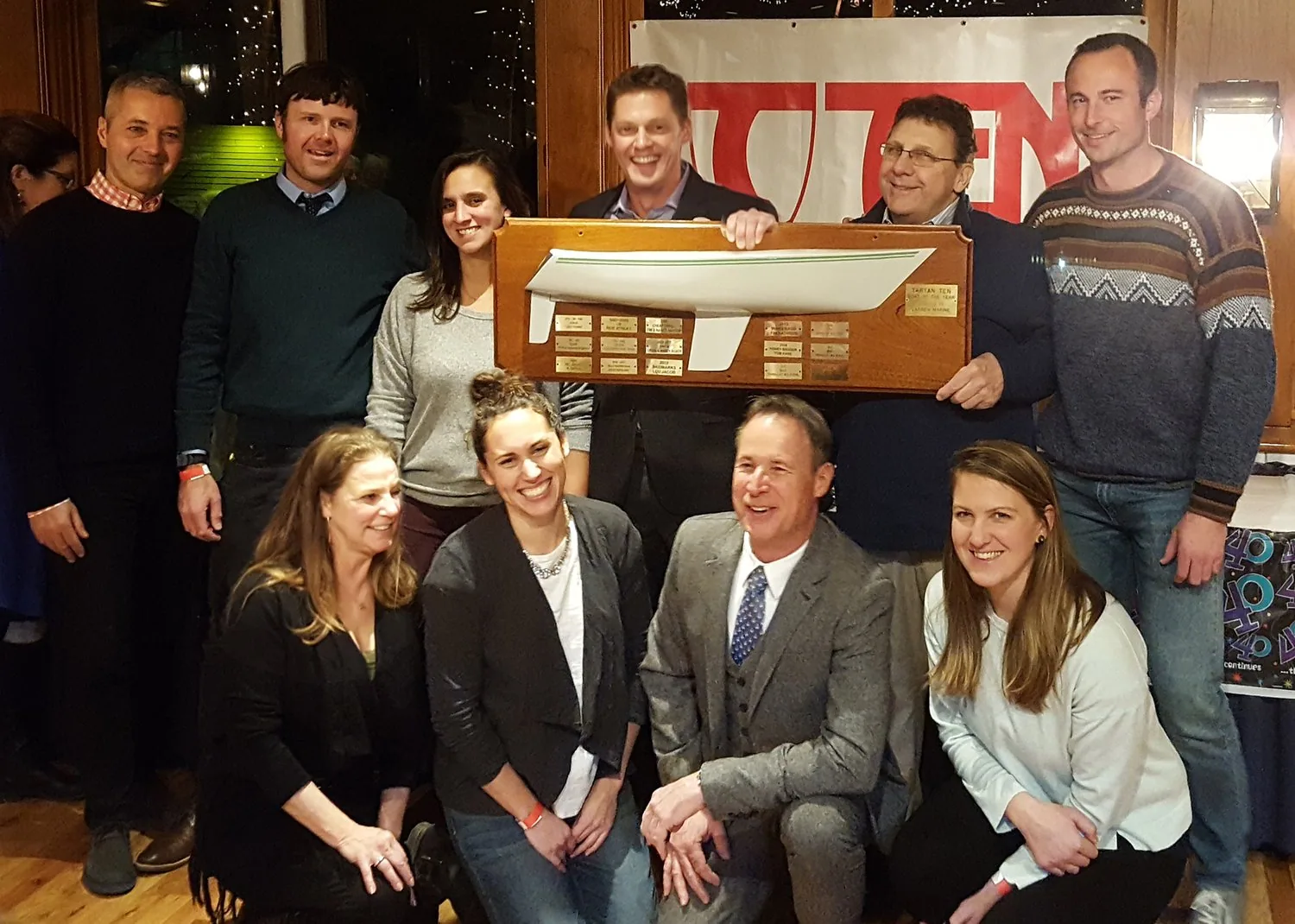 Ten people posing indoors with a Tartan 10 half-hull trophy plaque lined with engraved brass plates