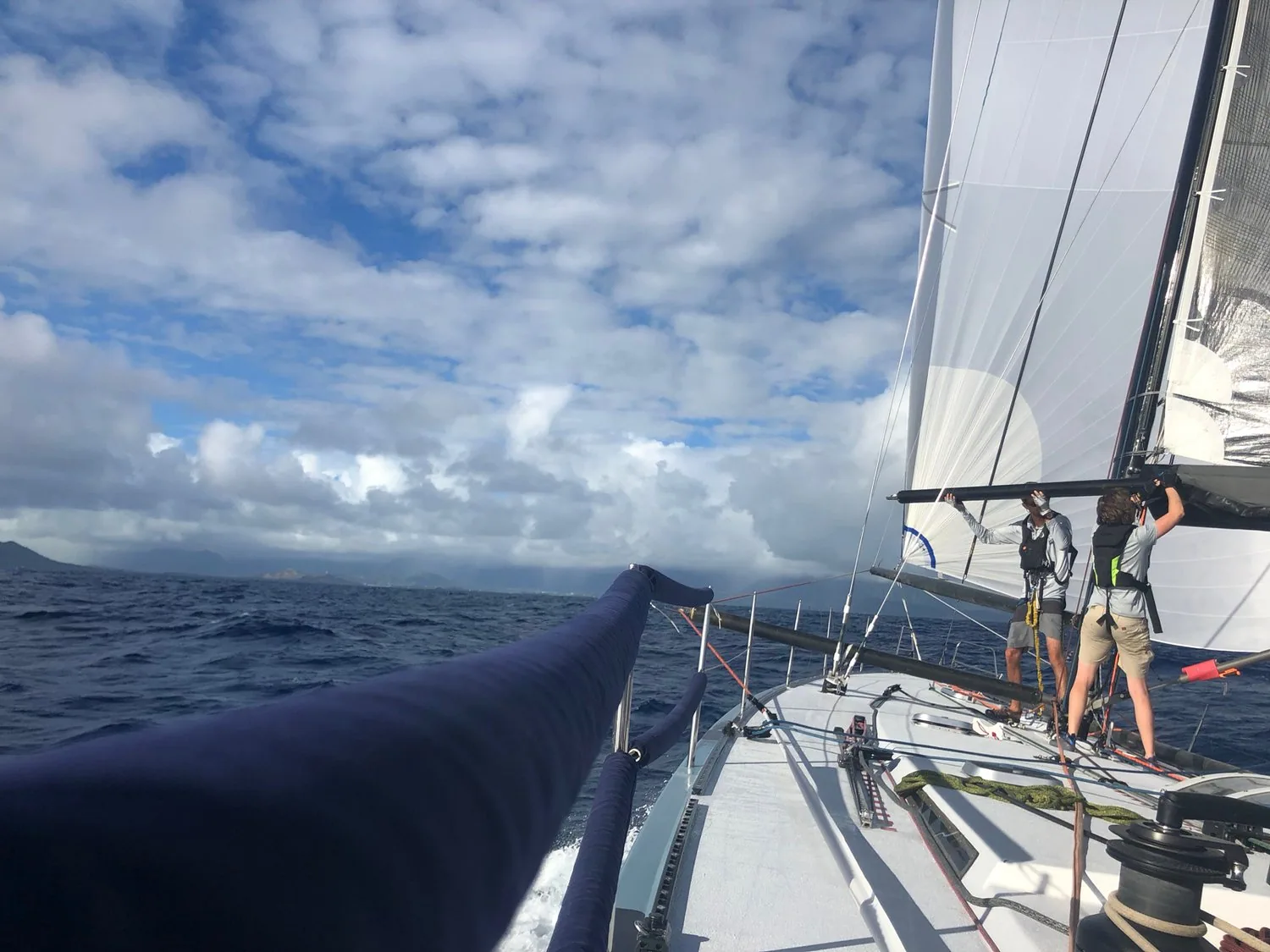 Looking forward along a dark blue sail cover on the boom toward two crew at the mast under white headsails, clouds ahead