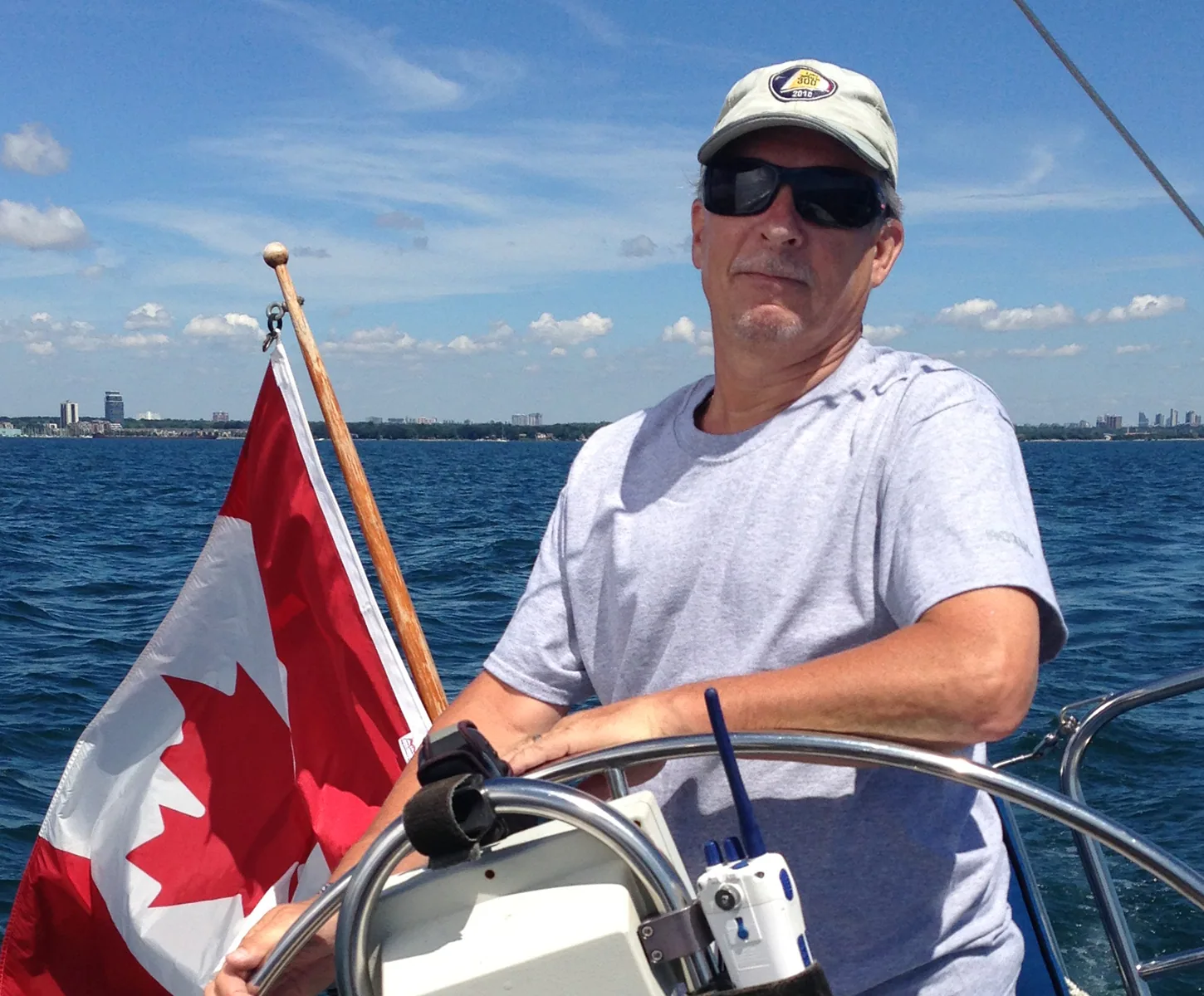 Randy St. Jacques at the wheel in a cap and sunglasses, Canadian flag flying off the stern, city skyline across the water