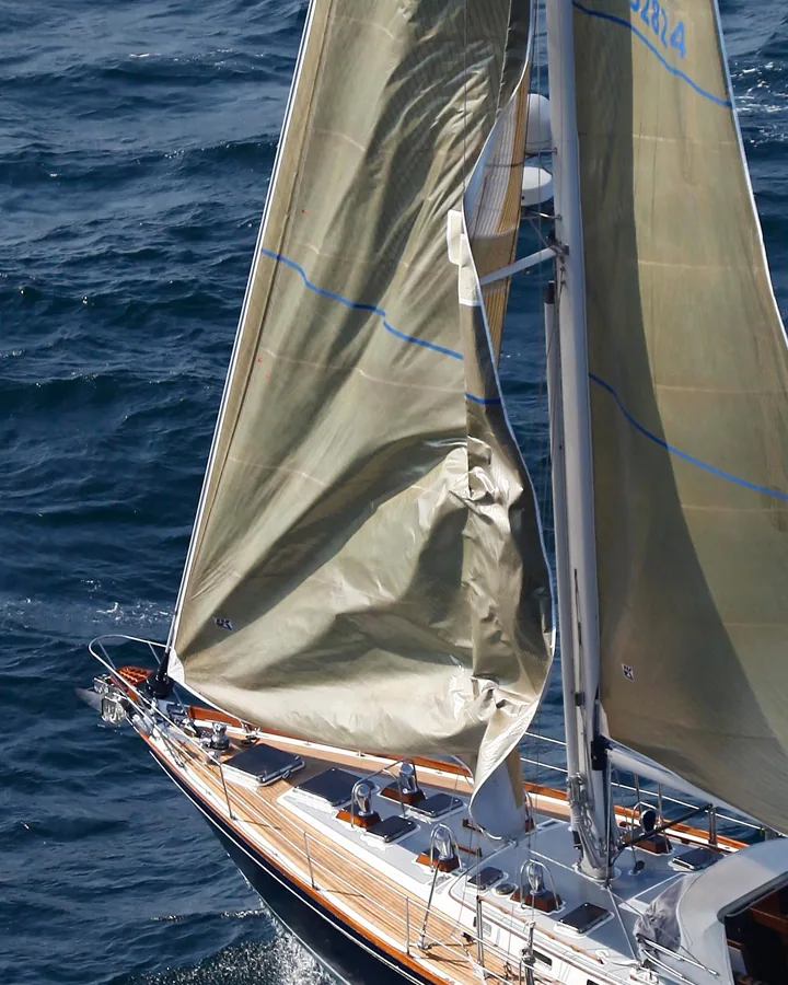 Tan laminate genoa dragging across the mast and spreaders during a tack, seen from aloft over a teak-decked yacht